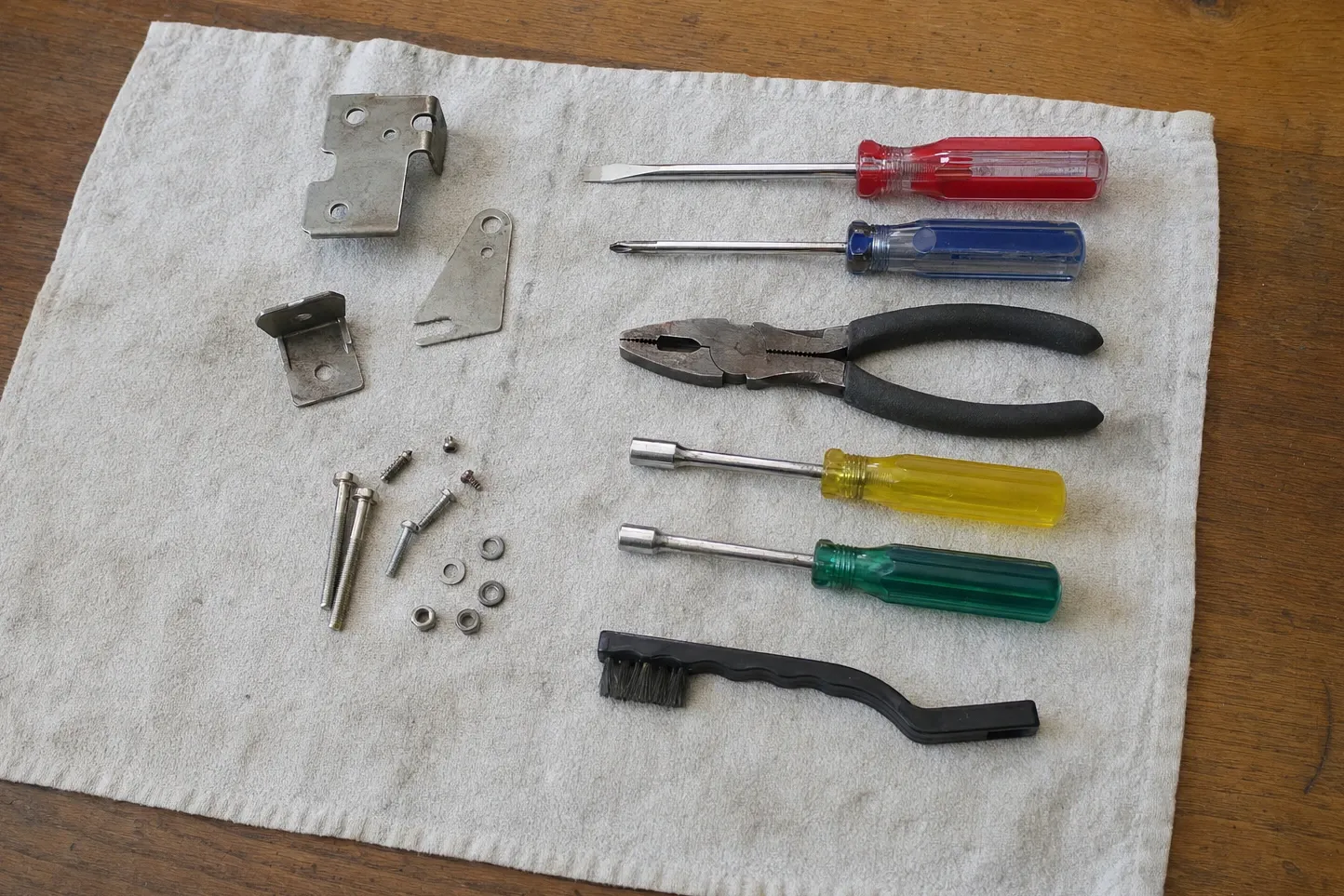 Stepped hinge bracket with an angled plate and an L-clip on a towel beside three long bolts, short screws, washers, nuts, two screwdrivers, pliers, nut drivers and a wire brush. Numbered marks are drawn over it, at the stepped hinge bracket, small angled plate and the long bolts and washers.